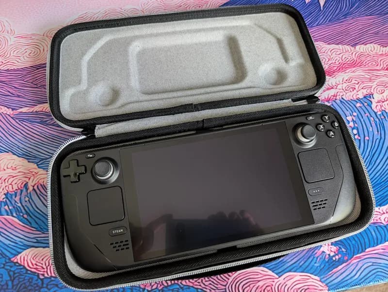 Source phone photo: a handheld game console in its open carrying case on a patterned desk mat
